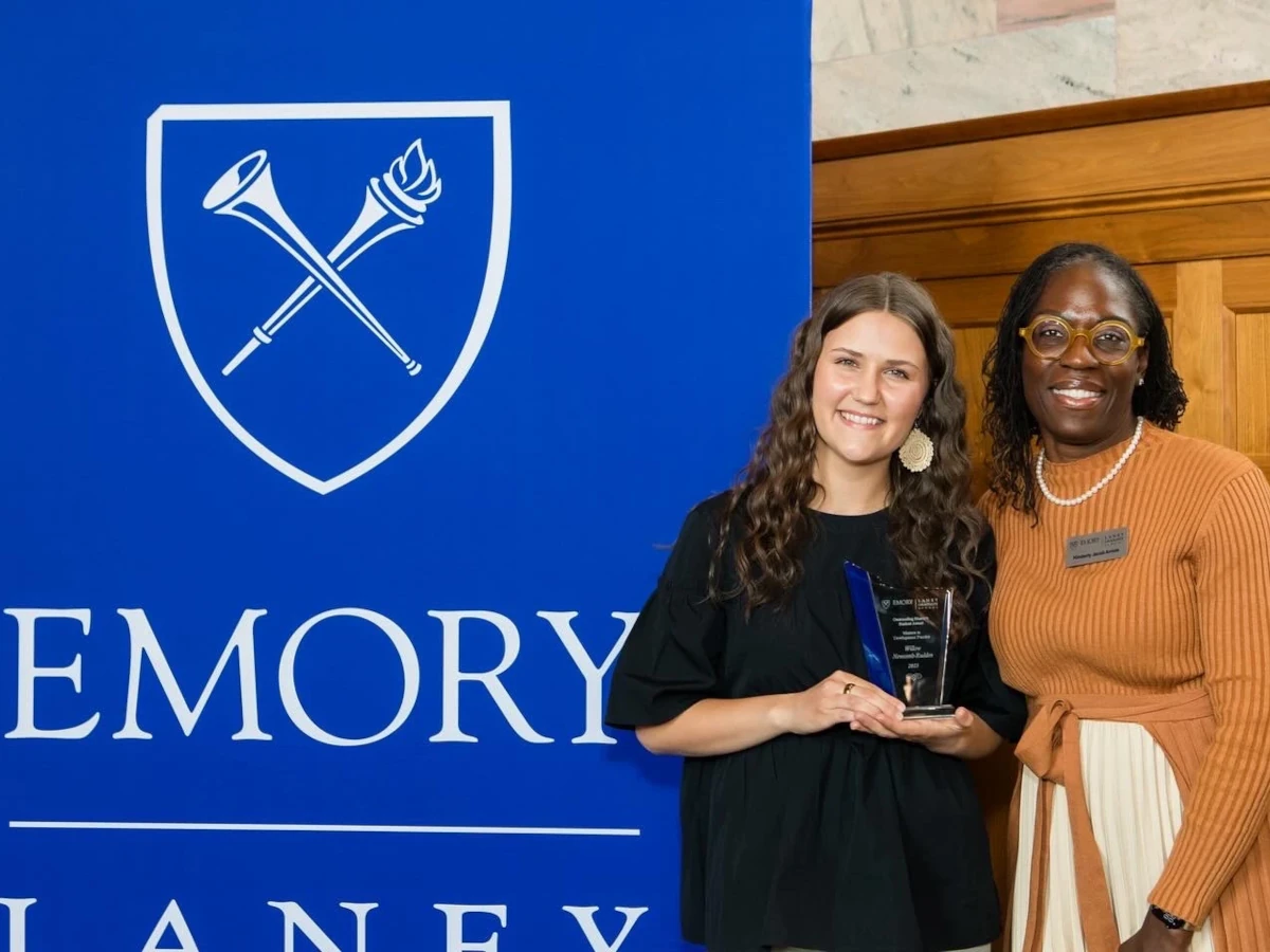 Student receiving an award at the Emory Laney Graduate School ceremony, standing with a faculty member in front of a blue Emory banner.